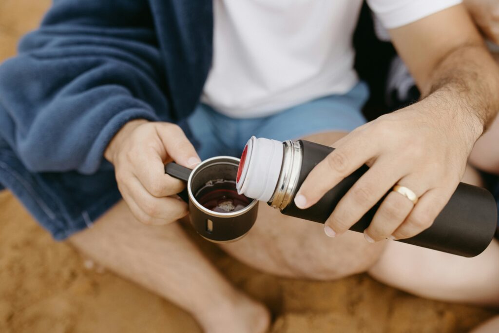 A man in a robe pours a drink from a thermos into a mug on a sandy beach.