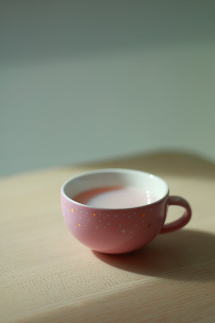 A soft pink mug with decorative stars, filled with a milky drink, sitting on a wooden surface in natural light.